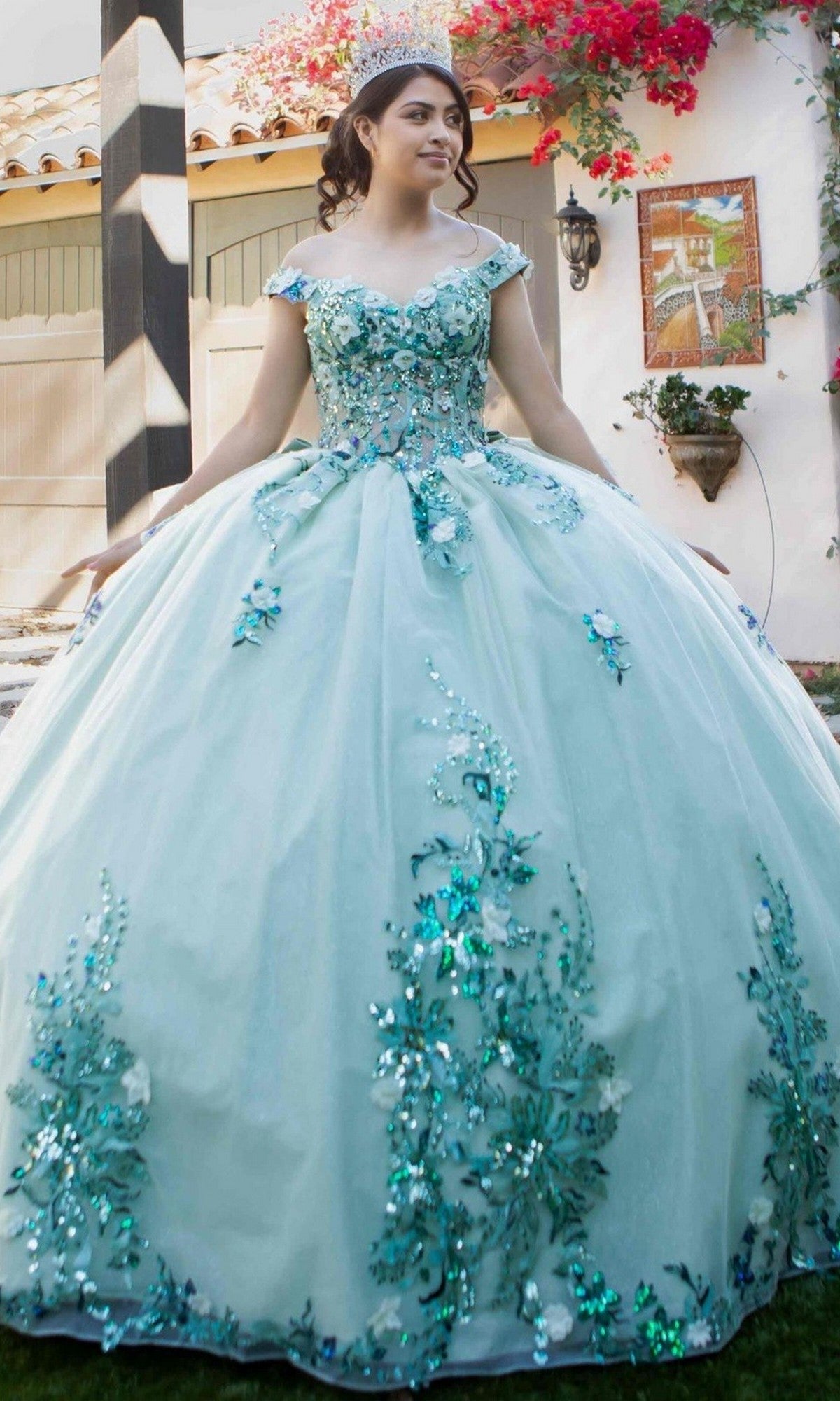A young woman smiles outdoors in the Quinceanera Ball Gown 8125J, an off-the-shoulder light blue dress with floral embellishments and a tiara on her head.